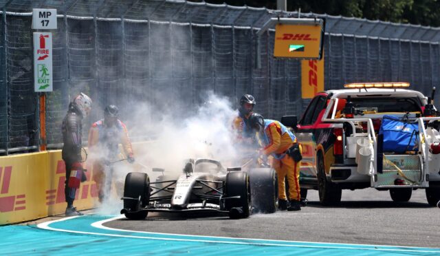 Formula 1 car halted after crash with smoke; safety crew and a fire extinguisher on-track near yellow barriers and a DHL sign above the fence.