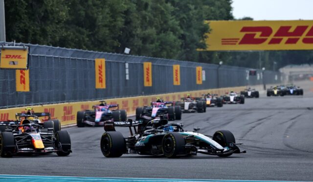 Multiple formula 1 cars race along a track, lined with DHL banners and yellow barriers on a straightaway.