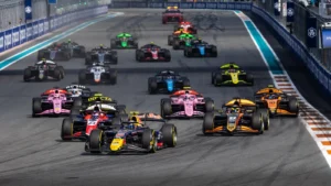 A pack of colorful Formula-style race cars accelerates from the starting line on a night race track.