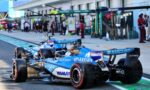 Blue Formula 1 race cars lined up in the pits with crew and garage buildings in the background, branding visible on the cars (Barclays, Komatsu).