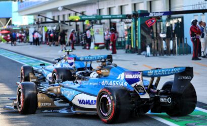 Blue Formula 1 race cars lined up in the pits with crew and garage buildings in the background, branding visible on the cars (Barclays, Komatsu).