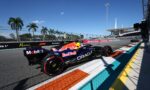Two Red Bull Formula 1 cars race side by side along a sunlit street circuit with colorful painted runoff areas, palm trees, and a grandstand in the background.