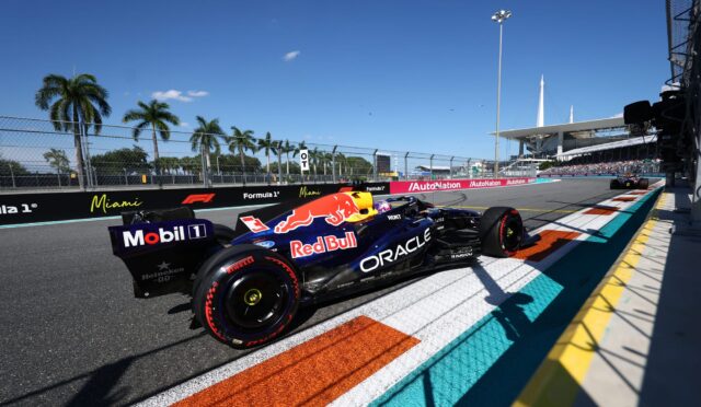 Two Red Bull Formula 1 cars race side by side along a sunlit street circuit with colorful painted runoff areas, palm trees, and a grandstand in the background.