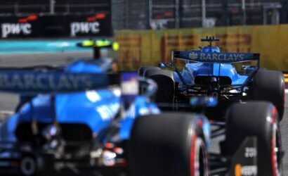Blue Formula 1 car centered on track with rear wing branded Barclays, racing with blur of other cars in foreground and background.