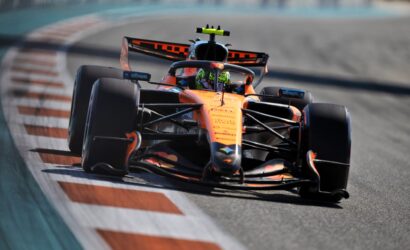 Orange Formula 1 race car speeds through a tight curve on a race track, driver wearing a neon helmet visible through the cockpit view