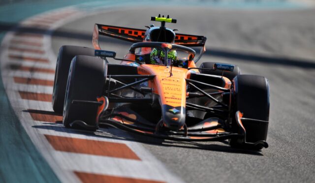Orange Formula 1 race car speeds through a tight curve on a race track, driver wearing a neon helmet visible through the cockpit view