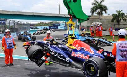 Pit stop scene with a Red Bull Formula 1 car in the foreground and orange-suited crew members nearby on a race track.",