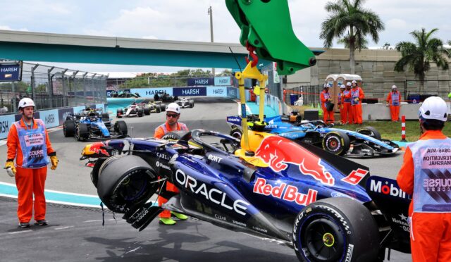 Pit stop scene with a Red Bull Formula 1 car in the foreground and orange-suited crew members nearby on a race track.",