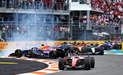 Formula 1 cars race through a tight turn as a large crowd watches from grandstands above the track, smoke rising from tires for effect of high-speed action.