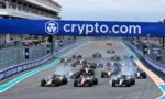 Pack of Formula 1 cars accelerating off the start line on a race track, with a large blue crypto.com banner overhead and a full grandstand crowd nearby.