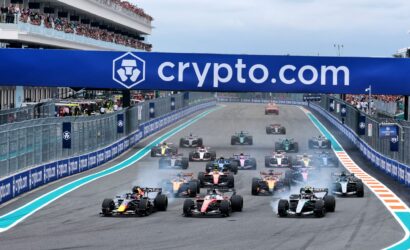 Pack of Formula 1 cars accelerating off the start line on a race track, with a large blue crypto.com banner overhead and a full grandstand crowd nearby.
