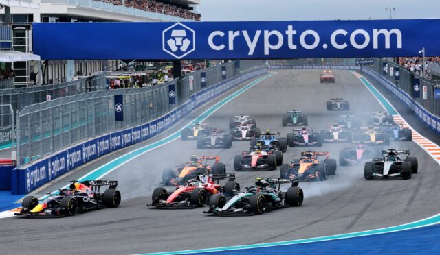 Formula 1 cars speeding off from the start on a blue-track circuit with sponsored barriers and a crowded grandstand above the track on crypto.com advertising.