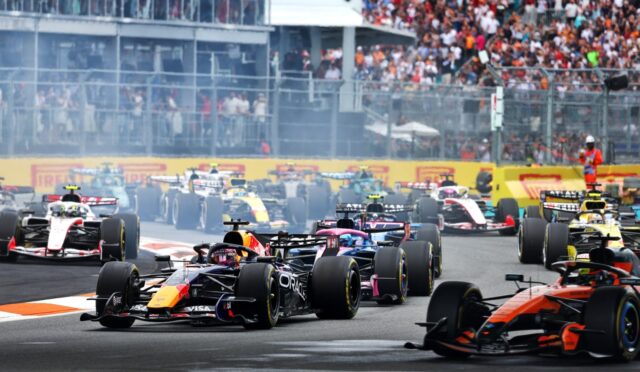 Multiple Formula 1 cars race off at the start on a city track with a large crowd in the stands behind a safety fence.
