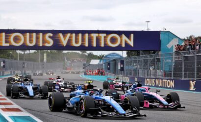 Group of Formula 1 cars at the race start beneath a Louis Vuitton banner, racing down the track with spectators along the barriers.