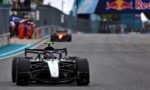 Front-on shot of a Mercedes Formula 1 car on the track, with pit crew and a Red Bull banner in the background.