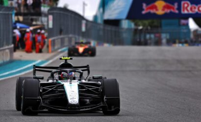Front-on shot of a Mercedes Formula 1 car on the track, with pit crew and a Red Bull banner in the background.
