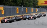 Row of Formula 1 race cars lined up on a straight track at the start of a race, DHL banners along the barrier behind them.