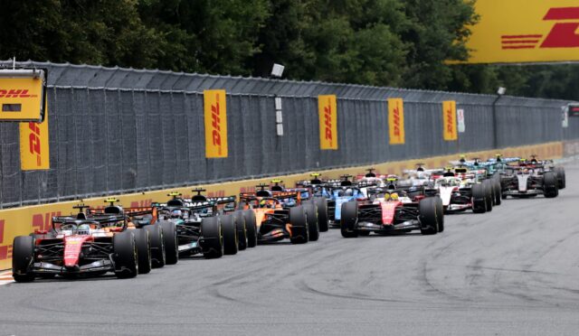Row of Formula 1 race cars lined up on a straight track at the start of a race, DHL banners along the barrier behind them.