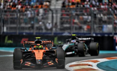 Orange Formula 1 car leads a race on a track, with a blurred crowd in the grandstands behind it.