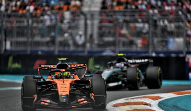 Orange Formula 1 car leads a race on a track, with a blurred crowd in the grandstands behind it.
