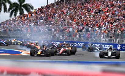 Formula 1 cars race side-by-side on a street circuit as a large crowd watches from grandstands behind a barrier, with smoke from tires and crypto.com advertising boards visible.