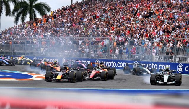 Formula 1 cars race side-by-side on a street circuit as a large crowd watches from grandstands behind a barrier, with smoke from tires and crypto.com advertising boards visible.