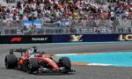 Red Formula 1 race car speeding on a track with a packed grandstand behind a fence and Formula 1 branding in the barriers on a sunny day.