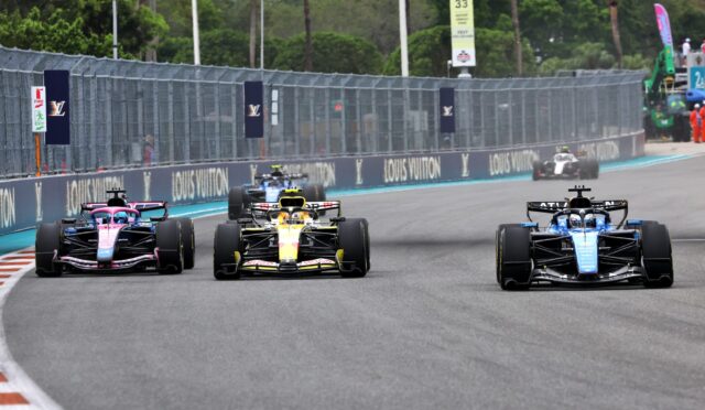 Four Formula 1 cars race side by side on a track with Louis Vuitton banners along the barrier wall.