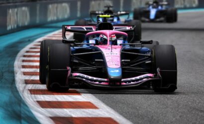 Front-on shot of a pink-blue Formula 1 car leading on a race track, with a second car visible in the blurred background behind it.