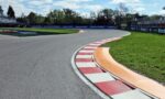 Wide view of a race track corner with red-and-white curbs, green grass, sponsor banners, and empty grandstands under a blue sky.