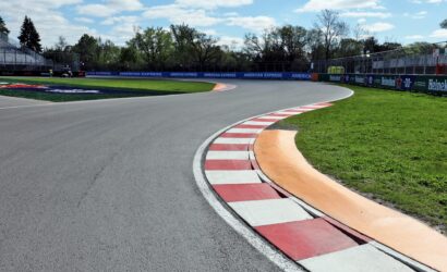 Wide view of a race track corner with red-and-white curbs, green grass, sponsor banners, and empty grandstands under a blue sky.