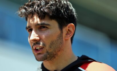 Close-up of a man with dark hair and stubble, looking to the right, wearing a red life jacket near blue water.