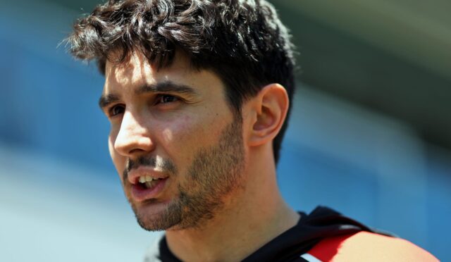 Close-up of a man with dark hair and stubble, looking to the right, wearing a red life jacket near blue water.