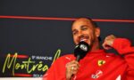 Man in red Ferrari jacket smiling while holding a microphone during an interview against a dark backdrop with red neon lines and sponsor logos nearby