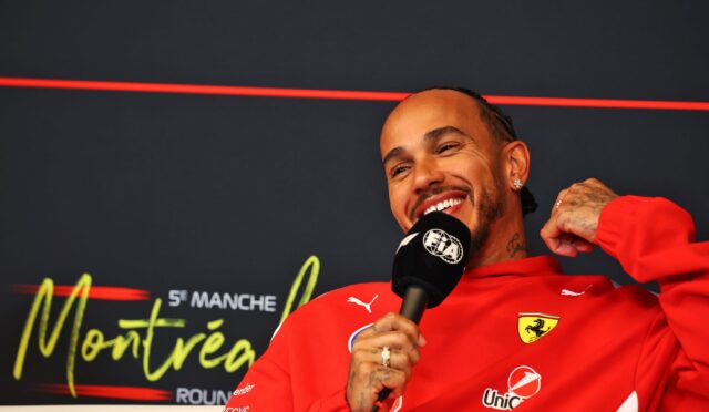 Man in red Ferrari jacket smiling while holding a microphone during an interview against a dark backdrop with red neon lines and sponsor logos nearby