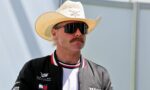 Man wearing a white cowboy hat and large black sunglasses, looking to the side, in a black jacket with logos visible on the chest and sleeve.