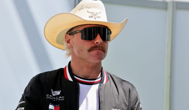 Man wearing a white cowboy hat and large black sunglasses, looking to the side, in a black jacket with logos visible on the chest and sleeve.