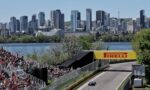 Racing car on a track approaching the turn with large audience stands and a city skyline in the background under a clear blue sky; Pirelli signage visible.