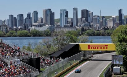 Racing car on a track approaching the turn with large audience stands and a city skyline in the background under a clear blue sky; Pirelli signage visible.