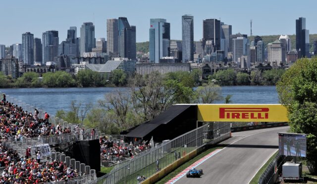 Racing car on a track approaching the turn with large audience stands and a city skyline in the background under a clear blue sky; Pirelli signage visible.