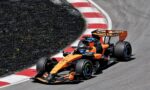 Two orange Formula 1 cars race through a tight turn, hugging a red-and-white barrier on the track edge