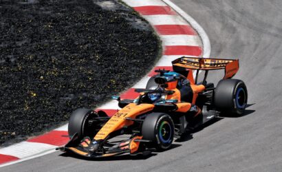 Two orange Formula 1 cars race through a tight turn, hugging a red-and-white barrier on the track edge
