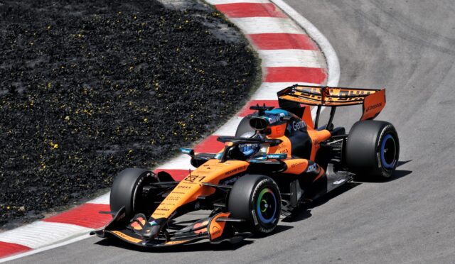 Two orange Formula 1 cars race through a tight turn, hugging a red-and-white barrier on the track edge