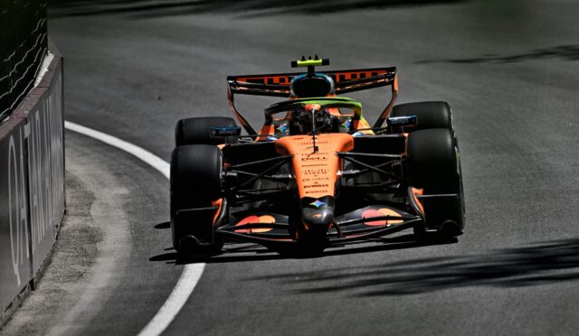 Orange Formula 1 car speeds along a curved race track, barrier on the left and the driver visible in the cockpit.