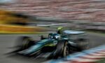 Motion-blurred green Formula 1 car speeding along a race track with red-and-white barriers and a blurred grandstand in the background.