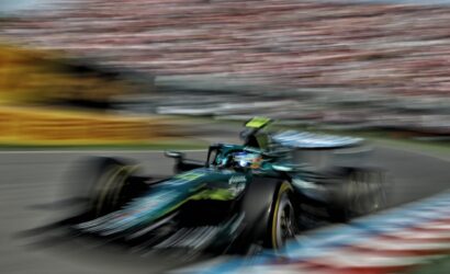 Motion-blurred green Formula 1 car speeding along a race track with red-and-white barriers and a blurred grandstand in the background.