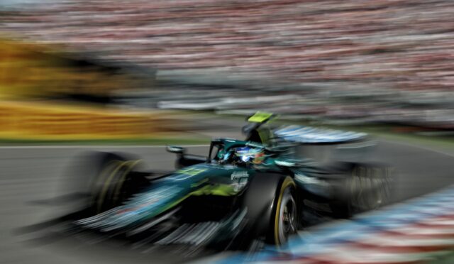 Motion-blurred green Formula 1 car speeding along a race track with red-and-white barriers and a blurred grandstand in the background.