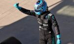 Race car driver in a blue helmet and black racing suit gives a thumbs-up, sponsor logos visible on the suit.