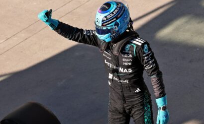 Race car driver in a blue helmet and black racing suit gives a thumbs-up, sponsor logos visible on the suit.