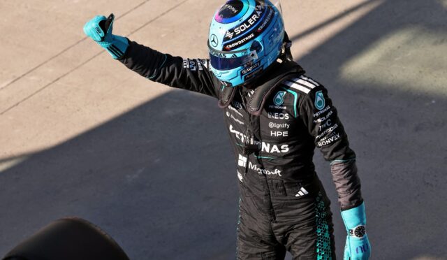 Race car driver in a blue helmet and black racing suit gives a thumbs-up, sponsor logos visible on the suit.